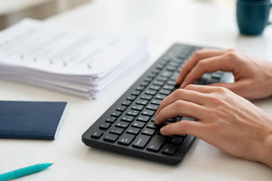 Hands typing on a keyboard next to a stack of documents and a passport