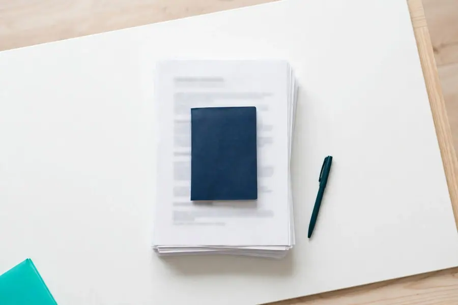 A closed passport on top of a stack of documents, with a pen beside it